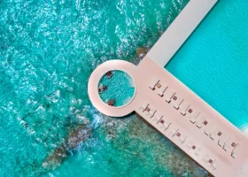 Vista aerea zenitale della piscina infinity a sfioro del NAKAI Alimathà Resort, isola di Alimathà, atollo di Vaavu, Maldive: il distintivo elemento architettonico circolare a forma di anello che si affaccia direttamente sull'Oceano Indiano, il camminamento fiancheggiato da lettini prendisole e le acque turchesi con la barriera corallina affiorante sul fondale.