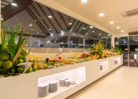 Buffet serale del ristorante principale del NAKAI Alimathà Resort, isola di Alimathà nell'atollo di Vaavu, Maldive: lungo bancone bianco con nicchie luminose e piatti impilati, ampia scelta di insalate, piatti freddi e frutta fresca, scenografica composizione di noci di cocco, stelle marine e foglie tropicali, sotto una copertura vetrata con tetto in legno tipico della formula all inclusive.