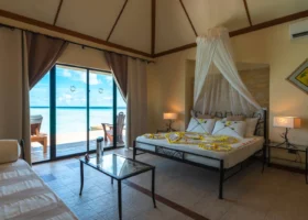 Interno di una Water Villa overwater del NAKAI Alimathà Resort sull'isola di Alimathà, atollo di Vaavu, Maldive: letto matrimoniale con baldacchino in velo di zanzariera e decorazione floreale a cuore, lampade brandizzate NAKAI, soffitto a padiglione e ampie vetrate scorrevoli che si aprono sulla terrazza privata con sdraio e vista diretta sulla laguna e sull'Oceano Indiano.