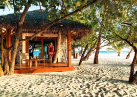 Beach Bungalow con tetto in makuti e pareti in corallo, affacciato sulla spiaggia di sabbia bianca dell'isola di Dhiggiri, nell'atollo di Vaavu alle Maldive; ampia veranda in legno con sedute, palme da cocco e ombra tropicale, ospite in piedi sull'ingresso e laguna dell'Oceano Indiano sullo sfondo nella luce dorata del tardo pomeriggio.