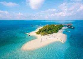 Veduta aerea dell'intera isola di Dhiggiri, nell'atollo di Vaavu alle Maldive, coperta di fitta vegetazione tropicale e palme, circondata da un ampio sandbank di sabbia bianca e dalle acque turchesi e blu dell'Oceano Indiano; sulla destra il pontile con le over water villa, cielo sereno con nuvole leggere e barriera corallina visibile in trasparenza sotto la superficie.