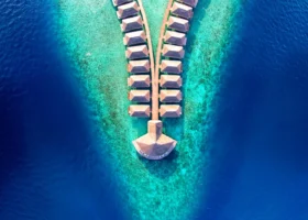 Ripresa aerea zenitale con drone delle over water villa disposte in doppia fila simmetrica lungo il pontile in legno dell'isola di Dhiggiri, nell'atollo di Vaavu alle Maldive; i tetti a punta si stagliano sopra la barriera corallina e i gradienti di colore dell'Oceano Indiano, dal turchese chiaro sul reef al blu profondo delle acque circostanti.