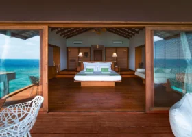 Interno di una over water villa dell'isola di Dhiggiri, nell'atollo di Vaavu alle Maldive, con letto king size e cuscini verdi al centro, pavimento e soffitto a travi in legno scuro, pareti vetrate scorrevoli aperte sulla veranda e vista diretta sull'acqua turchese dell'Oceano Indiano; poltroncina in metallo intrecciato bianco in primo piano sul ponte esterno.