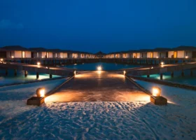 Pontile in legno illuminato da lanterne calde all'ora blu, con le over water villa allineate simmetricamente sopra la laguna dell'isola di Dhiggiri, nell'atollo di Vaavu alle Maldive; sabbia bianca in primo piano, riflessi dorati sull'acqua e cielo notturno blu intenso, atmosfera romantica ideale per una cena di coppia sull'Oceano Indiano.