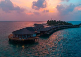 Veduta aerea al tramonto delle over water villa con tetto in makuti disposte lungo il pontile curvo sopra la laguna dell'isola di Dhiggiri, nell'atollo di Vaavu alle Maldive; cielo colorato di rosa e arancio riflesso sull'acqua calma dell'Oceano Indiano, con la vegetazione dell'isola e le luci calde delle villa in lontananza.