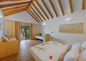 Interno luminoso di un beach bungalow con alto soffitto a capriata in travi di legno chiaro a vista, letto matrimoniale con decorazione floreale di ibisco e scritta di benvenuto, pavimento in parquet e ampia vetrata aperta sulla vegetazione, al NAKAI Maayafushi Resort, atollo di Ari Nord (Alif Alif), Maldive.