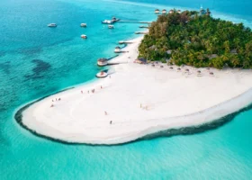 Lingua di sabbia bianca e finissima che si protende nella laguna turchese trasparente, con ospiti in spiaggia, lettini, ombrelloni in makuti e pontili sullo sfondo dell'isola di Maayafushi, atollo di Ari Nord (Alif Alif), Maldive, Oceano Indiano.
