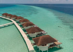 Fila di over water bungalow con tetti in tegole marroni allineati lungo il pontile sopraelevato sulla laguna cristallina dai fondali sabbiosi e coralli affioranti, al NAKAI Maayafushi Resort, atollo di Ari Nord (Alif Alif), Maldive, Oceano Indiano.