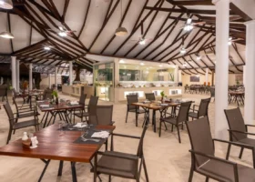 Sala interna del ristorante principale con pavimento in sabbia bianca, soffitto a capriate in legno scuro, ventilatori a pale, tavoli in legno apparecchiati e isola buffet centrale illuminata, al NAKAI Maayafushi Resort, atollo di Ari Nord (Alif Alif), Maldive.