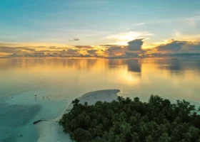 Tramonto dorato sull'oceano calmo con il sole e le nuvole riflessi sulla superficie della laguna e la lingua di sabbia dell'isola di Maayafushi in primo piano, incorniciata dalle palme da cocco, atollo di Ari Nord (Alif Alif), Maldive, Oceano Indiano.