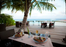 Cena romantica in riva al mare al Niva Kuramathi Maldives, con tavolo per due sotto una palma, aragosta alla griglia, calici di champagne e lanterne con candele, pontile in legno e laguna cristallina sullo sfondo, Isola di Kuramathi, Atollo di Rasdhoo (Ari Nord), Maldive, Oceano Indiano.