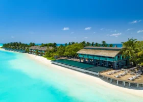 Vista aerea della spiaggia del Niva Kuramathi Maldives con lunga piscina a sfioro affiancata al ristorante principale dal tetto curvo in makuti, lettini bianchi sul pontile in legno e ville sulla spiaggia allineate lungo la sabbia bianca, laguna turchese dell'Isola di Kuramathi, Atollo di Rasdhoo (Ari Nord), Maldive, Oceano Indiano.