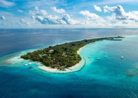 Vista aerea dall'alto dell'intera isola tropicale del Niva Kuramathi Maldives, allungata e ricoperta di vegetazione lussureggiante, circondata dalla laguna turchese con imbarcazioni all'ancora e dal blu profondo dell'Oceano Indiano, Atollo di Rasdhoo (Ari Nord), Maldive.