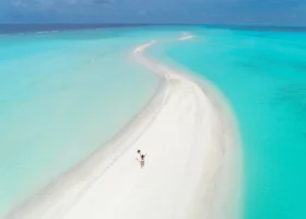 Ripresa con drone di una lunga lingua di sabbia bianchissima che si incurva nella laguna turchese del Niva Kuramathi Maldives, con una persona a braccia alzate al centro e la barriera corallina visibile sotto le acque cristalline, Isola di Kuramathi, Atollo di Rasdhoo (Ari Nord), Maldive, Oceano Indiano.