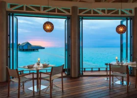 Interno del ristorante overwater del Niva Kuramathi Maldives al crepuscolo, con tavoli in legno apparecchiati con calici, lampade a sospensione in rattan e ampie vetrate a soffietto aperte sull'Oceano Indiano e su una water villa sotto il cielo rosato del tramonto, Isola di Kuramathi, Atollo di Rasdhoo (Ari Nord), Maldive.