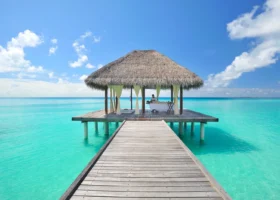 Padiglione spa overwater del Niva Kuramathi Maldives con tetto in makuti al termine di un pontile in legno, lettino da massaggio e tende bianche affacciate sulla laguna turchese e cristallina sotto un cielo azzurro con nuvole, Isola di Kuramathi, Atollo di Rasdhoo (Ari Nord), Maldive, Oceano Indiano.
