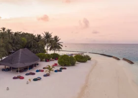 Spiaggia del Niva Kuramathi Maldives al tramonto con cielo rosa e pesca, area lounge con pouf colorati sotto un padiglione dal tetto in makuti, palme curve e sabbia bianca lambita dalla laguna calma, Isola di Kuramathi, Atollo di Rasdhoo (Ari Nord), Maldive, Oceano Indiano.