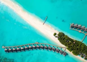 Ripresa aerea dall'alto delle ville overwater del Niva Kuramathi Maldives disposte in due file curve sopra la laguna turchese, con lingua di sabbia bianca, pontile in legno e vegetazione tropicale al centro dell'isola, Isola di Kuramathi, Atollo di Rasdhoo (Ari Nord), Maldive, Oceano Indiano.
