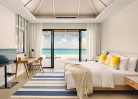 Interno luminoso di una Beach Villa con letto king size, cuscini gialli, tappeto a righe blu e ampie vetrate scorrevoli aperte sulla spiaggia di sabbia bianca e sulla laguna turchese dell'isola di Vakarufalhi, nell'Atollo di Ari Sud, Maldive.