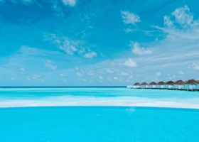 Piscina a sfioro dalle acque azzurre che si fonde con la laguna e con il cielo, con la fila di ville sull'acqua dal tetto in makuti sullo sfondo, presso il resort Nova Maldives sull'isola di Vakarufalhi, Atollo di Ari Sud, Maldive.