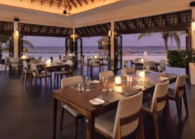 Sala ristorante open air al tramonto con tavoli in legno scuro, sedie imbottite, candele accese e tetto in makuti, affacciata sulla piscina e sull'Oceano Indiano tra le palme, presso Nova Maldives sull'isola di Vakarufalhi, Atollo di Ari Sud, Maldive.