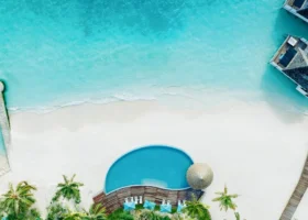 Ripresa aerea dall'alto della spiaggia di sabbia bianchissima con piccola piscina circondata da palme e ville dal tetto in paglia, lambita dalla laguna turchese e affiancata dalle ville sull'acqua dell'isola di Vakarufalhi, Atollo di Ari Sud, Maldive.