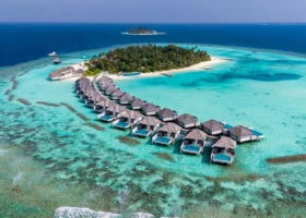 Ripresa con drone della lunga fila di ville sull'acqua dal tetto in makuti, ciascuna con piscina privata, disposte ad arco sulla laguna turchese davanti all'isola ricca di palme di Vakarufalhi, Atollo di Ari Sud, Maldive.