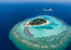 Veduta aerea dell'intera isola di Vakarufalhi con la vegetazione tropicale al centro e la fila curva di ville sull'acqua affacciata sulla barriera corallina, circondata dalle acque turchesi e dal blu profondo dell'Oceano Indiano nell'Atollo di Ari Sud, Maldive.