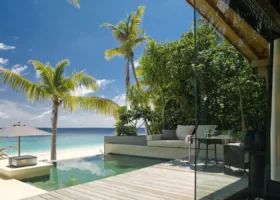 Terrazza in legno di una beach villa con piscina privata a sfioro affacciata sulla spiaggia di sabbia bianca; daybed imbottito, ombrellone e coppa di champagne davanti alla vetrata scorrevole, palme da cocco inclinate sul mare turchese sull'isola di Hadahaa, atollo di Gaafu Alifu, Maldive, Oceano Indiano.