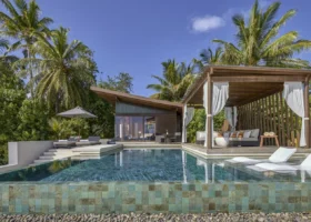 Esterno di una pool villa con ampia piscina rivestita in mosaico verde-oro, cabana in legno con tende bianche svolazzanti e daybed, lettini bianchi semisommersi e villa dal tetto spiovente immersa nella vegetazione tropicale sull'isola di Hadahaa, atollo di Gaafu Alifu, Maldive.