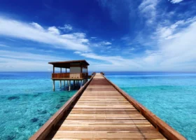 Pontile in legno che conduce in prospettiva verso un padiglione overwater su palafitte, sospeso sull'acqua cristallina che sfuma dal turchese al blu sopra il reef corallino, sotto un cielo azzurro con nuvole leggere sull'isola di Hadahaa, atollo di Gaafu Alifu, Maldive, Oceano Indiano.