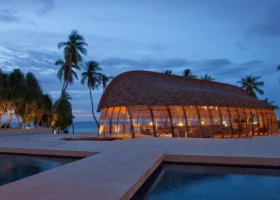 Padiglione ristorante illuminato al tramonto con l'iconico tetto curvo in scandole a forma di dhoni rovesciato e struttura a costole in legno, riflesso nelle piscine a specchio in primo piano tra palme da cocco stagliate contro il cielo blu del crepuscolo sull'isola di Hadahaa, atollo di Gaafu Alifu, Maldive.