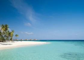 Spiaggia di sabbia bianca finissima lambita da una laguna turchese digradante, con palme da cocco chine sull'acqua a sinistra e una fila di ville overwater visibili all'orizzonte sotto un ampio cielo azzurro sull'isola di Hadahaa, atollo di Gaafu Alifu, Maldive, Oceano Indiano.