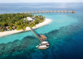 Vista aerea obliqua del pontile d'arrivo in legno con un dhoni tradizionale maldiviano ormeggiato sul reef, la spiaggia bianca a mezzaluna, le beach villa immerse nella vegetazione e la fila di ville overwater che si estende sull'oceano dell'atollo di Gaafu Alifu, Maldive.