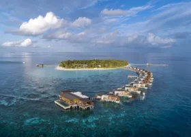 Vista aerea della lunga fila curva di water villa overwater con piscine private, sospese sul reef corallino visibile in trasparenza, che abbraccia l'isola boscosa e la spiaggia bianca sotto un cielo con nuvole cumuliformi sull'atollo di Gaafu Alifu, Maldive, Oceano Indiano.
