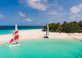 Due catamarani a vela, uno con vela rossa e bianca e uno con vela verde e blu, ormeggiati su una lingua di sabbia bianca lambita dalla laguna turchese, con un ospite pronto a salpare e la vegetazione tropicale dell'isola sullo sfondo, al Reethi Faru Resort nell'Atollo di Raa, Maldive, Oceano Indiano.