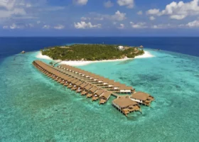 Vista aerea ravvicinata della lunga fila di ville overwater con tetti in makuti collegate da pontili sopra il reef corallino e la laguna turchese, con l'isola di Filaidhoo ricoperta di palme sullo sfondo, al Reethi Faru Resort nell'Atollo di Raa, Maldive, Oceano Indiano.