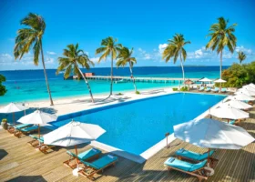 Piscina a sfioro dall'acqua azzurra con deck in legno, lettini con cuscini turchesi e ombrelloni bianchi, affacciata sulla spiaggia bianca, il pontile e la laguna cristallina incorniciata dalle palme, al Reethi Faru Resort sull'isola di Filaidhoo, Atollo di Raa, Maldive, Oceano Indiano.