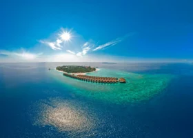Vista aerea dell'isola di Filaidhoo con la lunga fila curva di ville overwater dai tetti in makuti sospese sulla laguna turchese, il reef corallino affiorante e il sole a raggiera sul cielo blu, presso il Reethi Faru Resort nell'Atollo di Raa, Maldive, Oceano Indiano.