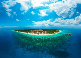 Panoramica dall'alto in drone dell'intera isola di Filaidhoo circondata dall'anello di reef corallino e dalla laguna turchese, con vegetazione tropicale, edifici centrali dai tetti in makuti e nuvole cariche sull'Oceano Indiano, al Reethi Faru Resort nell'Atollo di Raa, Maldive.