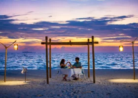Cena romantica per due sulla spiaggia al tramonto, coppia che brinda con calici di vino sotto una pergola in bambù illuminata da lanterne artigianali su pali di legno, tavolo con tovaglia bianca e secchiello per lo champagne sulla sabbia bianca; sullo sfondo il cielo violaceo e arancione si specchia sull'oceano di Bathala, nell'Atollo di Ari Nord alle Maldive, con un isolotto all'orizzonte.