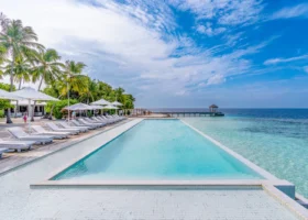 Piscina a sfioro rettangolare con bordo in mosaico bianco affacciata sull'oceano, fiancheggiata da una lunga fila di lettini prendisole bianchi e ombrelloni, palme da cocco sulla sinistra e un pontile con gazebo che si allunga sul mare; l'acqua azzurra della piscina si fonde visivamente con la laguna di Bathala, nell'Atollo di Ari Nord alle Maldive, sotto un cielo terso dell'Oceano Indiano.