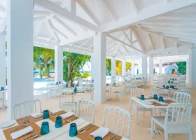 Ristorante open air con pavimento in sabbia bianca, ampia struttura dal tetto bianco spiovente con travi e capriate a vista e colonne candide, tavoli bianchi apparecchiati con bicchieri turchesi, tovagliette in bambù e posate; le aperture laterali incorniciano palme e vegetazione tropicale con la zona buffet sullo sfondo, nel cuore del resort sull'isola di Bathala, Atollo di Ari Nord, Maldive.