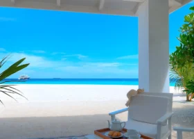 Veranda coperta di un beach bungalow con tetto bianco spiovente e colonna candida, poltrona in legno bianco con cappello di paglia e tavolino con vassoio in legno, teiera in porcellana, due tazze e croissant; oltre il deck in legno si apre la spiaggia di sabbia corallina bianca e la laguna turchese di Bathala, nell'Atollo di Ari Nord alle Maldive, con uno yacht all'orizzonte nell'Oceano Indiano.