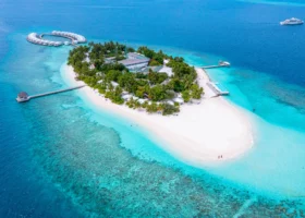 Veduta aerea da drone dell'intera isola corallina di forma allungata, ricoperta di fitta vegetazione tropicale e palme, con la fila curva di water villa collegate da pontile sulla sinistra, piscina, jetty e spiaggia bianca circondata da lagune turchesi e reef corallino; l'isola di Bathala emerge dall'Oceano Indiano nell'Atollo di Ari Nord alle Maldive, con uno yacht ancorato nel blu profondo.
