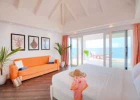 Interni luminosi di un water bungalow con alto soffitto bianco a travi a vista, letto king size con lenzuola bianche, cappello di paglia e pareo corallo, divano arancione con cuscini bianchi e borsa da spiaggia, palme in vaso e stampe marine incorniciate alle pareti; le vetrate scorrevoli con tende color corallo aprono sul deck privato con lettini affacciato sull'oceano di Bathala, nell'Atollo di Ari Nord alle Maldive.