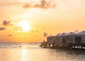 Fila di water bungalow bianchi su palafitte con tetti piramidali e ampi deck in legno, illuminati dalla luce calda e dorata del tramonto, con una figura in silhouette affacciata sull'acqua calma che riflette i toni aranciati del cielo; le ville overwater si stagliano sull'oceano dell'isola di Bathala, nell'Atollo di Ari Nord alle Maldive, nell'Oceano Indiano.
