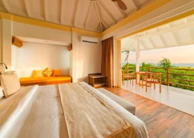 Interno di una beach house dal soffitto a volta bianco e pavimento in legno chiaro, con letto king, daybed dai cuscini arancioni e ampia vetrata aperta sulla veranda affacciata sul verde tropicale e sull'oceano nella luce dorata del tardo pomeriggio. Siyam World Maldives, Dhigurah, Atollo di Noonu, Maldive.