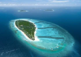 Veduta aerea dell'ampia isola di Dhigurah coperta di fitta vegetazione, circondata dalla barriera corallina e dalle lunghe file di water villa dal tetto verde che si protendono sulla laguna turchese; sullo sfondo altre isole dell'Atollo di Noonu nell'Oceano Indiano. Siyam World Maldives, Maldive.