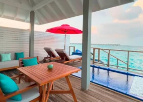 Deck privato in legno di una water villa con piscina a sfioro, ombrellone rosso, lettini e divano dai cuscini verdi e turchesi affacciati sulla laguna turchese; sullo sfondo la fila delle altre ville overwater sull'acqua. Siyam World Maldives, Dhigurah, Atollo di Noonu, Oceano Indiano.